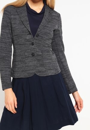 Woman wearing a fitted, dark grey buttoned blazer over a navy blue dress with a flared skirt, standing against a plain white background.