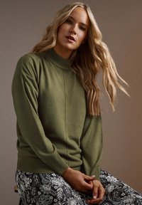 Maglione a maglia verde oliva con collo alto, spalle scese e polsini a coste, abbinato a pantaloni bianchi e neri con motivo floreale.