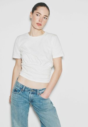 Kvinde med mørkt hår iført en hvid kortærmet top med rynker og blå jeans, stående med den ene hånd i lommen mod en ensfarvet hvid baggrund.