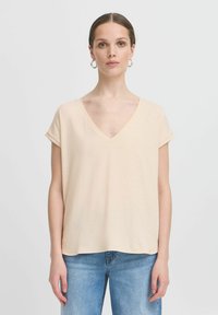 Žena svijetle puti i smeđe kose u niskoj punđi nosi srebrne naušnice u obliku obruča, bež majicu s V-izrezom i kratkim rukavima te plave traperice na bijeloj pozadini.