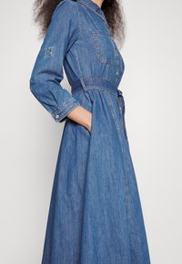 Denim shirt dress with a fitted waist, long sleeves, and side pockets. Light blue color, button-down front, and belted design for shape.