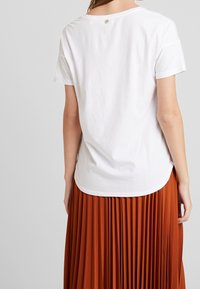 Camiseta blanca de manga corta con dobladillo redondeado, que presenta un pequeño detalle en la parte posterior. Combinada con una falda naranja con pliegues.