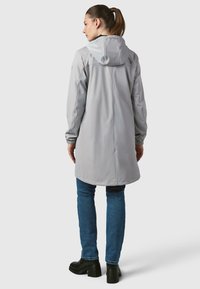 Lys grå regnjakke med hætte, lange ærmer og lige skæring, kombineret med blå jeans og kraftige sorte sko. Glat, vandafvisende stof.