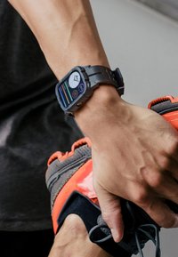 Digital sports watch with a black band and rectangular face displays time and activity stats; hand holds a black and red athletic shoe.