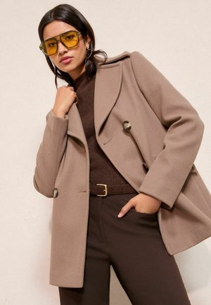 Brauner Wollmantel mit großen Knöpfen, getragen über einem dunkelbraunen Pullover und einer Hose. Das Modell trägt gelb getönte Brillen und präsentiert einen Lagenlook.