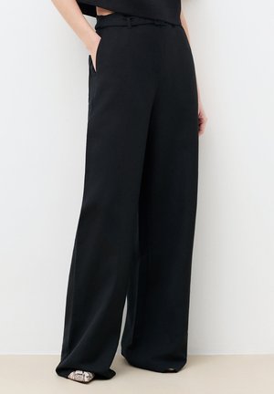 Kobieta nosząca czarne, szerokie spodnie z wysokim stanem i paskiem, ręce w kieszeniach, zestawione z butami w szpic o wzorze skóry węża.