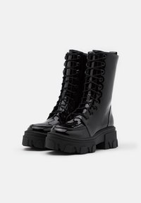 Bottes de combat noires brillantes avec semelles épaisses à crampons et devant à lacets, présentées côte à côte sur un fond blanc.