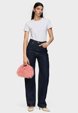 Mujer con camiseta blanca de manga corta, vaqueros oscuros de talle alto y pata ancha, tacones beige, sosteniendo un bolso rosa esponjoso.