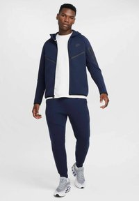 Mornarska zip-up kapuca z črnimi detajli, v kombinaciji z usklajenimi trenirkami. Nošena čez belo majico, dopolnjena s sivimi športnimi čevlji.