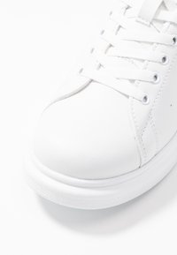 Bílá teniska s hladkým syntetickým svrškem, kulatou špičkou, šněrováním vpředu s textilními tkaničkami a texturovanou podrážkou pro lepší přilnavost.