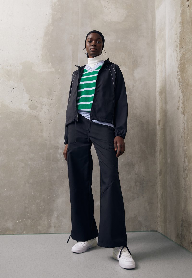 Chaqueta negra con cremallera, suéter a rayas verdes y blancas, pantalones anchos negros y zapatillas blancas. Tejido suave, diseño casual, acentos mínimos.