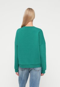 Zelená mikina s okrúhlym výstrihom, pružnými manžetami a lemom. Mäkký materiál a voľný strih, kombinovaná s svetlomodrými džínsami.