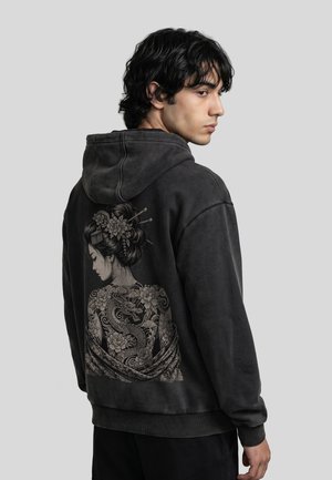 Jeune homme aux cheveux foncés portant un sweat à capuche noir avec une œuvre d'art style tatouage complexe représentant une femme avec des motifs floraux et de dragon.