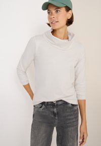 Hellgraues Oberteil mit Kapuze und langen Ärmeln, kombiniert mit dunkelgrauen Jeans. Der Stoff wirkt glatt mit enganliegenden Ärmeln und subtilen Nähdetails.
