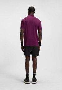 Dunkelviolettes Poloshirt mit kurzen Ärmeln und flachem Kragen, kombiniert mit schwarzen Shorts und Turnschuhen. Strukturierter Stoff mit glatter Oberfläche.