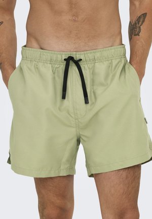 Uomo che indossa pantaloncini da bagno verde chiaro con coulisse nera, mani nelle tasche e tatuaggio di farfalla visibile sul braccio destro.