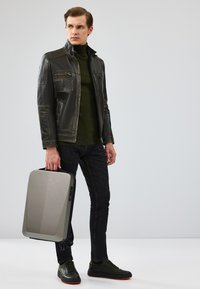 Veste en cuir noire pour hommes, col roulé vert foncé, jean foncé et baskets noires. Tenant une valise rigide gris argenté avec des accents.