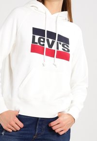 Sweat à capuche blanc avec une poche kangourou frontale, arborant un logo Levi's imprimé en noir, bleu marine et rayures rouges. Capuche à cordon de serrage et matériau doux.