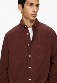 Ein junger Mann trägt ein knopfbares, dunkelrotes Cordhemd über einem weißen T-Shirt. Er blickt nach rechts mit einem neutralen Ausdruck.