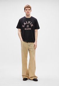 Črna bombažna majica s kratkimi rokavi z belim in roza grafičnim napisom "CALVIN KLEIN". Združena s svetlo rjavimi širokimi hlačami in črnimi čevlji. Enostavna ozadje.