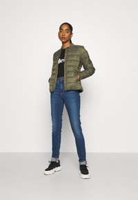 Olivgrüne, gesteppte Pufferjacke mit Stehkragen, zwei Seitentaschen, kombiniert mit blauen Jeans und schwarzen Turnschuhen.