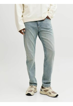 Moški, oblečen v svetlo modre kavbojke z ravnim krojem, kremno dolgo majico z dolgimi rokavi in bež superge, stoji z eno roko v žepu.