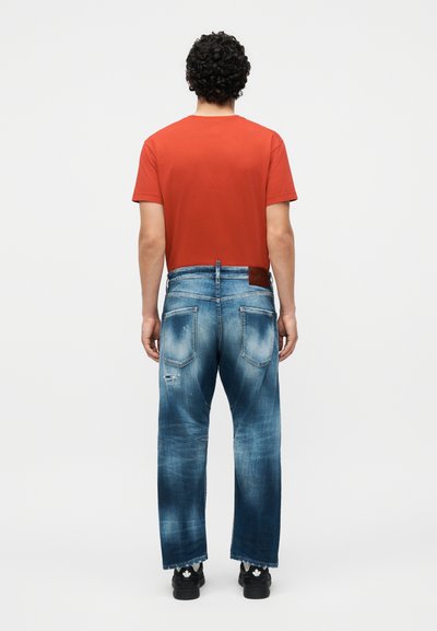 Άνδρας με την πλάτη, φοράει κόκκινο T-shirt, ξεθωριασμένο μπλε τζιν με φθαρμένες λεπτομέρειες και μαύρα αθλητικά παπούτσια με λευκά λογότυπα φύλλων.