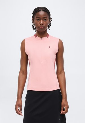 Kvinde med flettet hår iført en lyserød ærmeløs top med lynlås og sort nederdel, stående mod en ensfarvet lys baggrund.