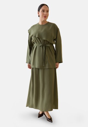 Donna con top verde oliva a maniche lunghe con cintura e gonna abbinata alla caviglia con scarpe scure marroni a punta, che guarda di lato.