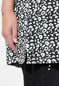 Zwarte stoffen top met witte abstracte vlekken, met een zijspli. Het kledingstuk is gecombineerd met donkere denim broeken met parelaccenten.