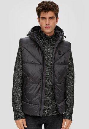 Gesteppteste schwarze Weste mit Kapuze, mit einem Reißverschluss ausgestattet. Wird über einem dunklen, strukturierten Strickpullover getragen. Logo auf der Brust.