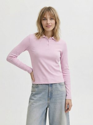 Ung kvinne med skulderlangt blondt hår iført en lys rosa langermet poloshorte og løse lyseblå jeans foran en ensfarget bakgrunn.