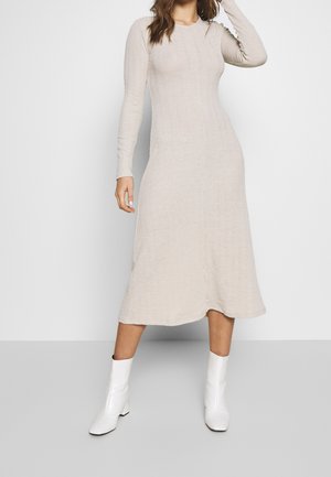 Femme portant une robe pull midi beige à manches longues, associée à des bottines blanches, debout devant un fond blanc uni.