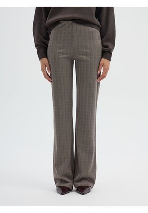 Donna che indossa pantaloni marroni a gamba larga con motivo a spina di pesce, maglione marrone scuro e scarpe bordò con punta.