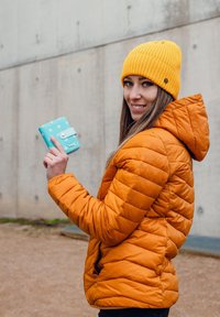 Woman in orange jacket and yellow beanie holding a blue polka-dot wallet, smiling, with a concrete wall in the background.