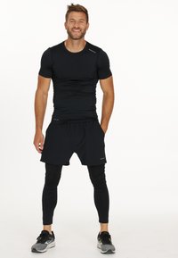 T-shirt et shorts de sport noirs portés sur des leggings noirs. L'ensemble présente un design ajusté et un matériau qui évacue l'humidité pour le confort.