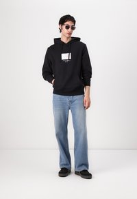 Felpa nera in cotone con grafica sul petto, abbinata a jeans di denim azzurri e scarpe nere. Il modello, indossando occhiali da sole, sta in piedi su uno sfondo neutro.