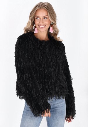 Vrouw in een zwart shaggy trui met lange mouwen en lichtblauwe jeans, glimlachend en naar de zijkant kijkend, met roze franjes oorbellen.