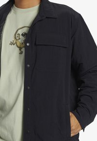 Dunkelmarineblaue Knopfjacke mit einer Brusttasche, strukturiertem Stoff und einem legeren Schnitt über einem hellgrünen T-Shirt mit einem Eidechsen-Design.