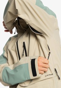 Giacca impermeabile beige e verde menta con polsini regolabili, molteplici tasche con zip e texture liscia. Il design presenta una vestibilità moderna e rilassata.