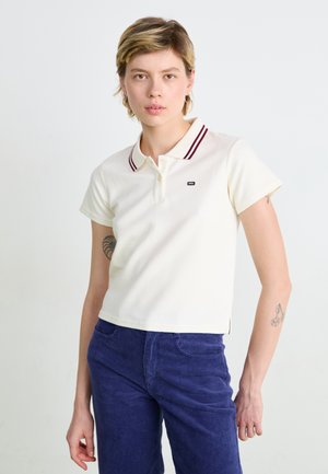 Polo in avorio con colletto a righe color mogano, maniche corte, texture a coste e un piccolo logo sul lato sinistro, abbinato a pantaloni di velluto a coste blu navy.