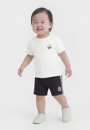 Väike laps naeratab, kannab valget t-särki ja musti lühikesi pükse lillemustriga, seisab ühevärvilise valge tausta ees.