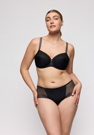 Vrouw poseert in zwart kanten beha met dubbele bandjes en bijpassende kanten zijden slip tegen een effen achtergrond.