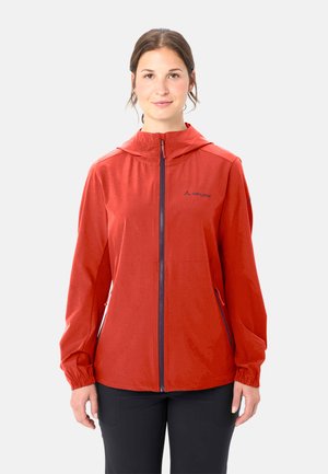 Vrouw die een rood jack met rits en capuchon en een zwarte broek draagt, staand tegen een effen lichte achtergrond, voorwaarts gericht met een neutrale uitdrukking.