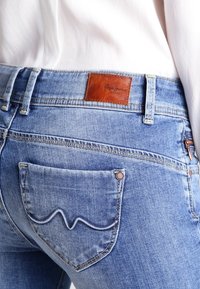 Jasnoniebieskie dżinsy z denimu o teksturowanym wykończeniu. Posiadają skórzaną metkę w kolorze brązowym na pasku oraz tylnią kieszeń z haftowanym wzorem fali.