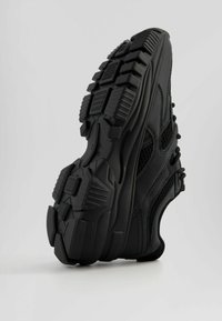 Czarne sportowe buty z teksturowaną gumową podeszwą, panelami z siateczki i zakrzywionym designem. Charakteryzują się wyraźnym wzorem bieżnika i wzmocnionymi akcentami.