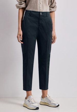 Person iført beige skjorte med knapper og optrukne ærmer, sorte korte cargobukser og hvide sneakers står mod en ensfarvet baggrund.