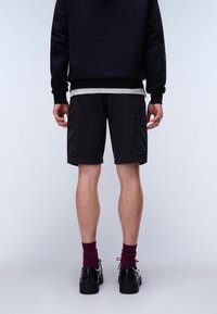 Isik, kes kannab musti lühikesi pükse, musti kingi ja burgundi punaseid sokke, seisab seljaga valge tausta ees.