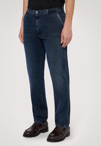 Tmavě modré džíny rovného střihu vyrobené z denimu, s knoflíkovým zapínáním, předními kapsami a jemným zesvětlením. Nohavice jsou kombinovány s hnědými botami.