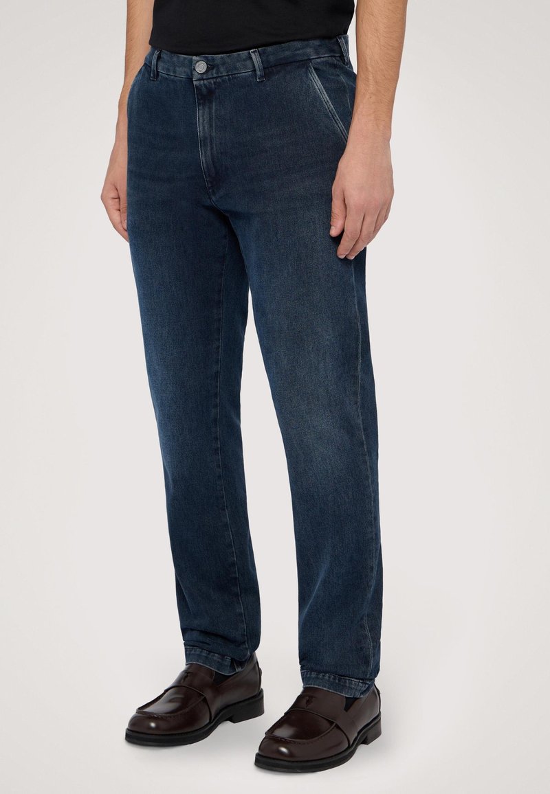 Tmavě modré džíny rovného střihu vyrobené z denimu, s knoflíkovým zapínáním, předními kapsami a jemným zesvětlením. Nohavice jsou kombinovány s hnědými botami.